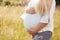 Cropped shot of unrecognizable young future mother being pregnant, keeps hands on tummy, stands on green field, wears white blouse