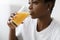 Cropped thirsty young pretty black lady in white t-shirt drinks fresh orange juice from glass indoor, profile