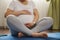 Cropped view of a expectant gravid woman holding her pregnant belly, sitting in lotus pose on blue exercise mat