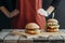 cropped view of girl in apron standing at wooden table with homemade cheeseburgers