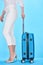 Cropped view of woman i white pants holding handle of blue colorful travel bag on blue background.