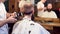 Cropped young barber haircutting with trimmer and brush, client sitting back in front of mirror with blurred reflection