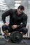 Crossfit heavy workout training by young bearded strong male in black hoodie sitting relaxing with iron barbell in bright sport