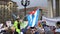 Crowd of protesters go. Tall guy in a green suit on stilts with flag.