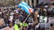 Crowd of protesters go. Tall guy in a green suit on stilts with flag.