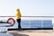 A cruise passenger in a yellow parka stands at the railing at the stern of a cruise ship and looks back at the blue open sea.