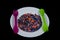 Crushed multicolored plastic pieces on white plate and green spoon and lilac fork .