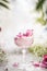 Crystal champagne glass with fancy pink drink with rose petal at white table with flowers and blurred green branches. Elegant
