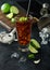 Cuba Libre cocktail in highball glass with ice and lime peel on bamboo stick with straw and fresh limes on black background