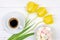 A Cup of black coffee on a saucer, a bouquet of yellow tulips and marshmallows on a white wooden background, top view