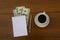 Cup of coffee, dollars, notepad and pen on wooden desk