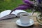 Cup of coffee, open book and bunch of purple summer flowers in glass vase on wooden table in the garden.