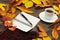 Cup of coffee, open notebook, pen and scarf among autumn leaves on wooden table