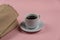 A cup of coffee and a paper bag against a pink background. Full espresso white cup with saucer. Sandwich, hamburger, or lunch in a