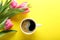 Cup of coffee and pink tulips on yellow background, top view
