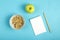 A cup with granola, a green apple and a notebook with a pencil to write calories on a blue background.