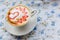 A Cup of hot cappuccino with a pattern of a red heart on the table with a tablecloth in a flower in a coffee shop. Close-up and bl