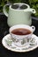 Cup of tea in a china teacup and saucer with a pastel green teapot reflected on a glass table