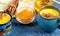 Cups of golden turmeric milk with cinnamon. Ingredients. Yellow napkin and blue backdrop. Close-up, selective focus