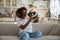 Curious excited young African woman holding modern VR glasses while sitting on sofa in living room