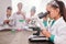 Curious schoolgirl in whitecoat looking in microscope by desk against classmates