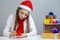 Curious Winsome Caucasian Female Girl in Santa Hat and White Shirt Holding Tiny Red Gift Box While Looking Inside Near Stack of