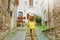 Curious young woman with yellow dress and hat goes upstairs in medieval street in Tuscany, Italy. Rear view of happy cheerful girl