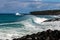 Curling wave trailing sea spray breaking near shore on Hawaiian cost. Strip of land in background with rocks and trees. Cloudy Sky