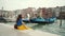 Curly brunette girl in colorful clothes sits on pier with gondola on Grand Canal