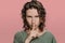 Curly caucasian young woman looks serious, holding finger at her lips asking shut up