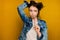 A curly dark-haired girl in a jeans stands on a yellow background, looks to the side, pressing the earpiece of glasses