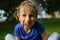 Curly girl with light brown hair is sitting on the grass. She holds a purple clover flower in her mouth. The baby smiles