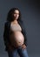 Curly haired pregnant woman with naker belly, wearing beige bra, gray blazer and blue jeans, thoughtfully looking aside