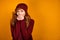 A curly red-haired girl in a burgundy sweater, scarf and hat stands on an orange background and warms her hands