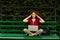 A curly young woman with closed eyes, working on a laptop, seated on a green bench in park touche her temples.