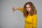 curly young woman in yellow clothes shows thumb down on gray background