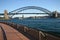 Curved embankment waterfront promenade with iconic Sydney Harbor Bridge. Urban coastal cityscape with historic landmark.