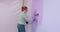 Curvy blonde woman paints wall with roller in light purple