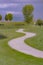 Curvy pathway small pond bench and trees on a grassy terrain under cloudy sky