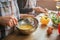 Cut view of man an woman cooking at table in kitchen. She blend eggs in bowl. He cut vegetables on desk. Glass of wine