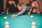 Cut view of young man looking concentrated. He aiming into billiard ball. Two slim models stand behind him.