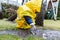 Cute adorable caucasian toddler boy in bright yellow raincoat and wellies playing alone at dirt muddy puddle during cold