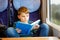 Cute adorable kid boy reading book during traveling by train. Happy child sitting near window. Family going on vacations