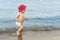 Cute adorable little blond caucasian serious pensive toddler boy in red panama walking through blue clear water waves of