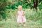 Cute adorable white Caucasian baby girl child in white dress standing in green summer park forest outside