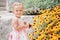 Cute adorable white Caucasian baby girl child in white dress standing among yellow flowers outside in garden park looking in