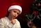 Cute angry girl in fur santa claus hat near the Christmas holiday tree thinking about the gift with grimacing face and looking se
