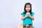 Cute Asian girl holds the lid of jar with coin and smiling looks at camera. Child put in lot of silver coins in jar.