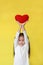 Cute asian little girl lift up red heart over her head isolated on yellow background