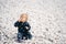 Cute baby in a blue overalls sits on a pebble beach, turning his head to the left and holding a pebble in his hand that